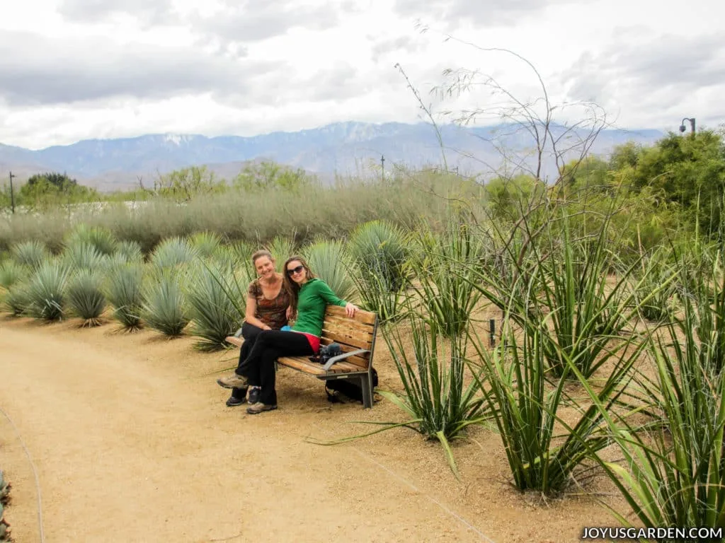 two women sitting on a bench in a desert garden with mountains in the background