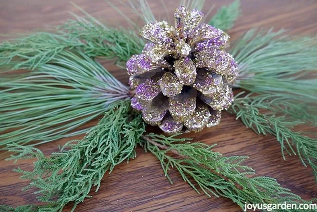 a pine cone glittered with gold & purple glitters sits on evergreen branches