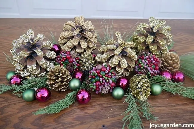 gold glittered pine cones, burgundy & green balls, & evergreen branches sit on a table