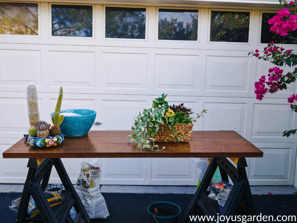 a work table outside a garage holds 2 dish gardens & a glazed turquoise pot