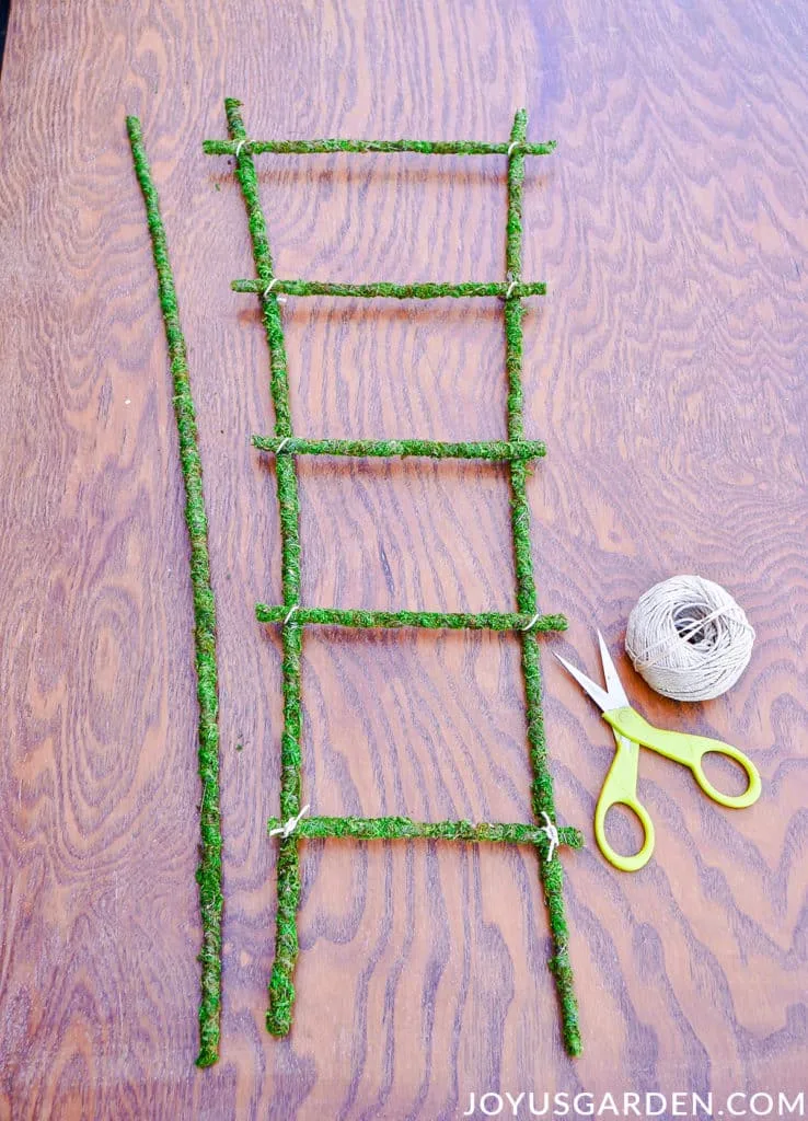 a homemade moss trellis sits on a work table next to a ball of twine & a pair of scissors